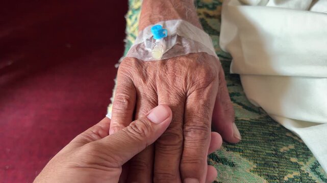 A comforting hand gently holds elderly hand with IV cannula secured by clear medical tape.