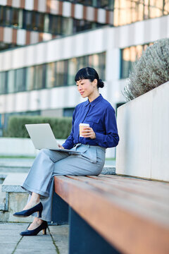Portrait of a young asian japanese businesswoman woman using a laptop computer and holding a cup of coffee  in a city park, surrounded by modern corporate office buildings