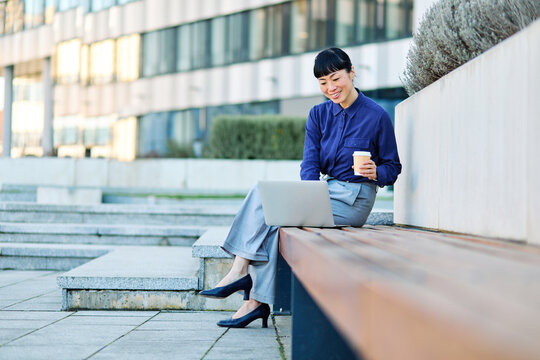 Portrait of a young asian japanese businesswoman woman using a laptop computer and holding a cup of coffee  in a city park, surrounded by modern corporate office buildings