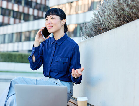 Portrait of a young asian japanese businesswoman woman using a smartphone mobile phone walking down the street, surrounded by moder corporate office buildings