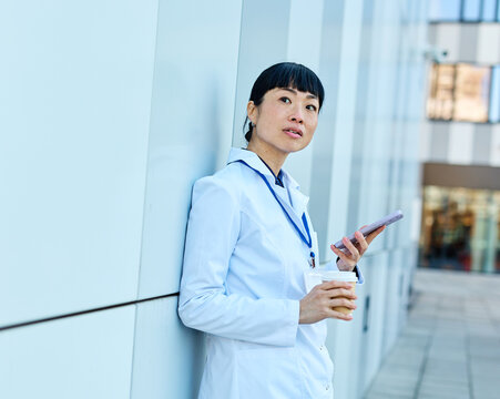 Portrait of an asian female woman doctor or nurse and  health care worker using a smartphone mobile phone holding a cup of coffee   in front of hospital or clinic outside
