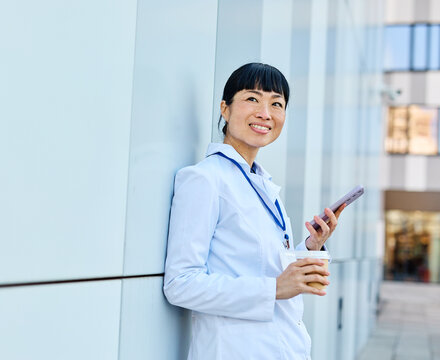 Portrait of an asian female woman doctor or nurse and  health care worker using a smartphone mobile phone holding a cup of coffee   in front of hospital or clinic outside