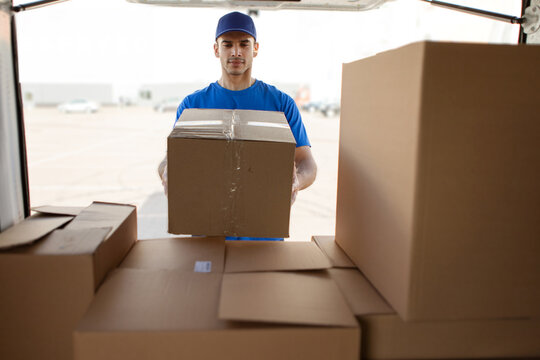 Man courier lifting large cardboard box out of van or loading parcels into van, preparing for delivery