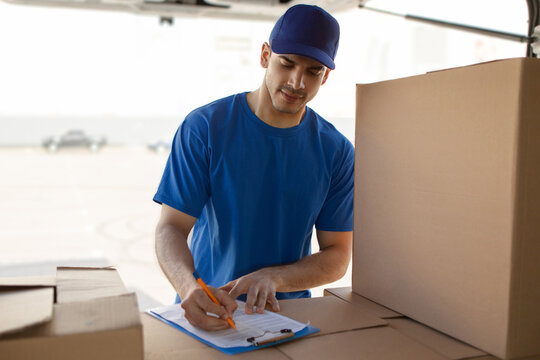 Delivery man filling documents while standing outdoors near van full of cardboard boxes, view from inside vehicle