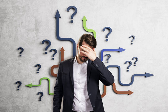 Confused businessman standing before many colorful question marks and arrows illustrating difficult choices and uncertainty on light background.