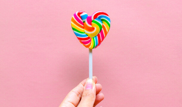 Hand holding colorful heart shaped lollipop isolated on pink background. Rainbow swirl candy with vibrant colors, sweet confectionery treat