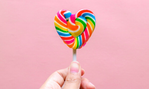 Hand holding colorful heart shaped lollipop isolated on pink background. Rainbow swirl candy with vibrant colors, sweet confectionery treat