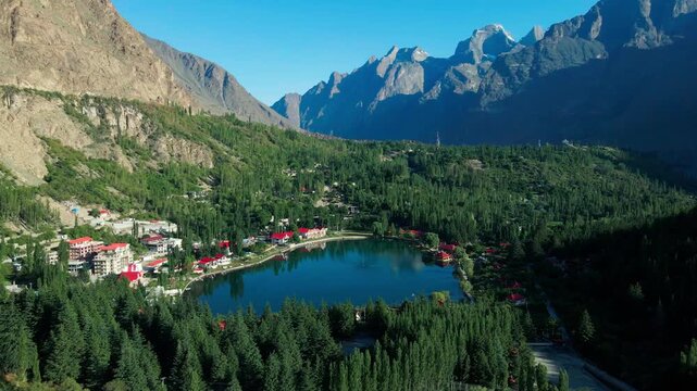 Aerial view of Shangrila Resort Skardu nestled among lush green trees and towering mountains by a calm lake in Skardu, GB, Pakistan.