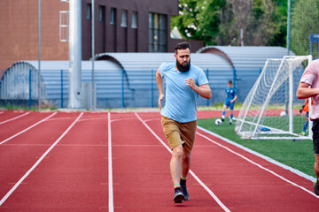 Fototapeta premium A man jogging on a red running track near a sports field, showcasing healthy exercise and outdoor activity with others playing in the background. A setting focused on activity and recreation.