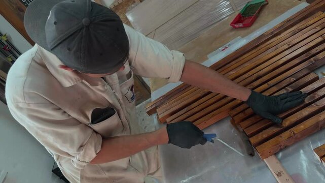 Dedicated male artisan in white uniform and cap applying dark wood stain to pine timber edges with roller in local home manufacturing workshop during furniture production medium dutch angle shot