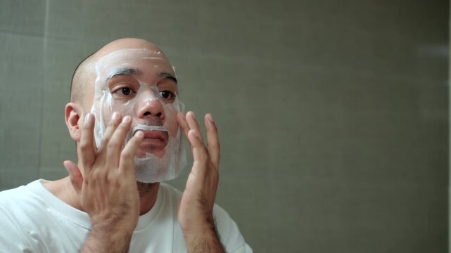 Man applying a sheet mask in front of a bathroom mirror during his morning skincare routine. Healthy aging longevity. Men self-care anti-aging skincare. wellness and preventive health concept.
