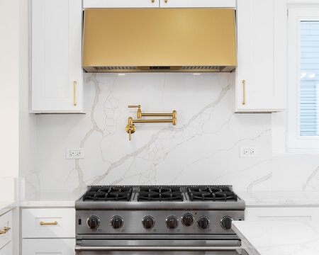 A white kitchen detail with a marble backsplash and countertop, stainless steel oven, and gold range hood, hardware, and pot filler faucet.