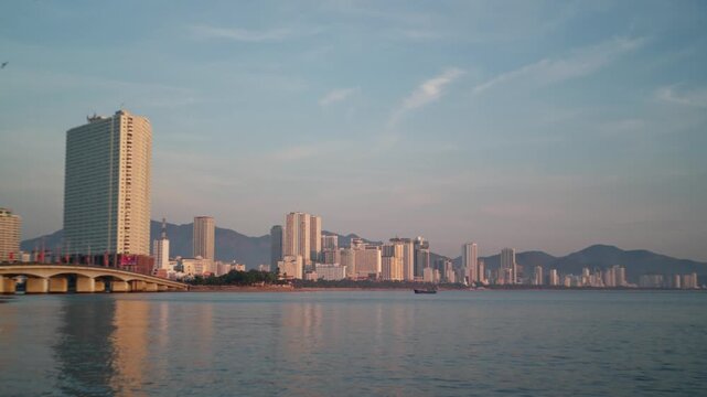 Coastal city skyline with tall waterfront towers, arched bridge and calm bay glowing in warm evening light, panoramic view