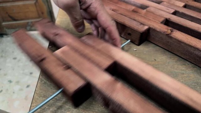 Close up of unrecognizable male hands threading stained wooden timber blocks onto a long metal threaded rod on a workbench during an authentic DIY furniture assembly process in private workshop