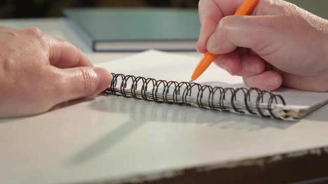 Hand writes with ballpoint pen in spiral-bound notebook on sheet of paper while sitting at table close-up