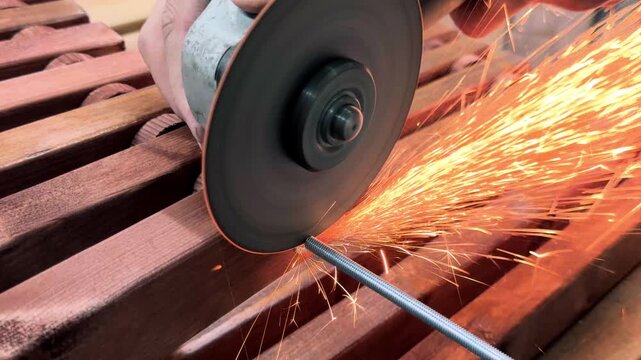 Handheld extreme close up of an unrecognizable self-made artisan trimming a steel threaded rod with a heavy angle grinder sending hot sparks flying during wooden furniture assembly in a garage shop