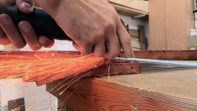 Handheld low angle shot of an anonymous self employed crafter using an angle grinder to cut a steel rod with flying sparks during wooden furniture assembly inside a local cottage industry workshop