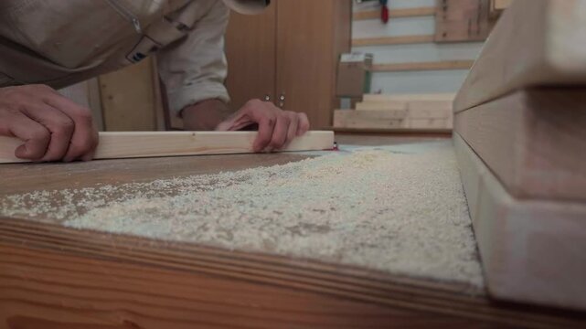 Macro view of anonymous woodworker hands carefully pushing timber piece on professional routing machine past blurred foreground stack of lumber covered with sawdust in realistic factory dolly shot