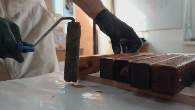 Skilled artisan in protective gloves applying rich dark liquid wood stain to raw pine timber edges with handheld roller in professional manufacturing workshop for custom furniture making project shot
