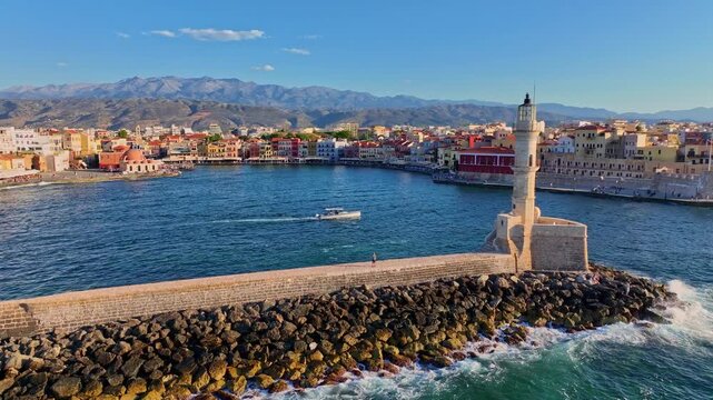 Aerial view of the historic Old Venetian Port of Chania with its iconic lighthouse, stone breakwater, and colorful waterfront buildings under a clear blue sky in Chania, Greece.