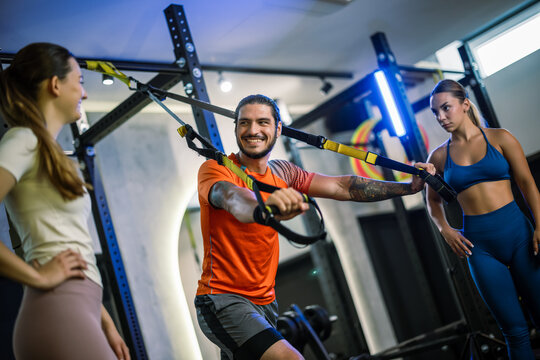 A personal trainer demonstrating a suspension strap exercise while two women watch in a gym