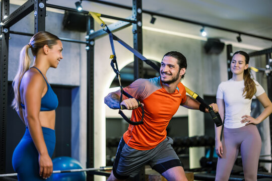 Standing fitness coach showing suspension strap technique to two female athletes at the gym