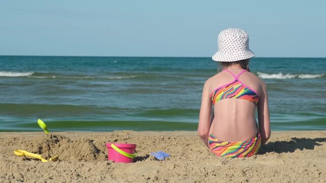 Girl in swimsuit and panama hat sitting on sandy beach looking at sea waves next to toys. Concept of childhood relaxation and summer vacation