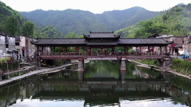 A traditional covered bridge spans a calm river, with mountain scenery and village structures in the background, with people walking on it.