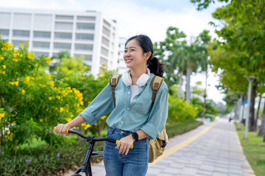 Asian woman smiles and wears headphones while riding a bicycle in a park.