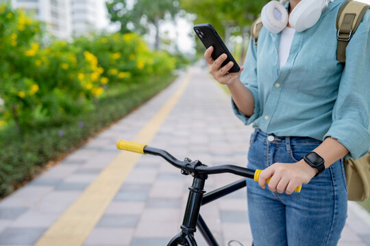 Happy young Asian woman, with a backpack and using her smartphone, stands by the roadside in a park, on her way to work at the office.