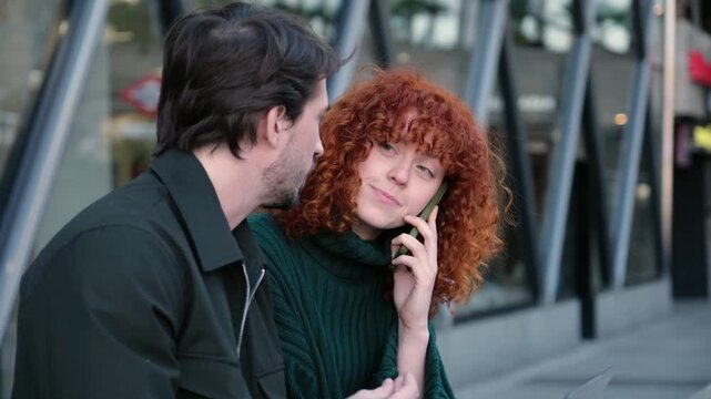 Man and woman working together on a laptop while the woman talks on the phone outdoors