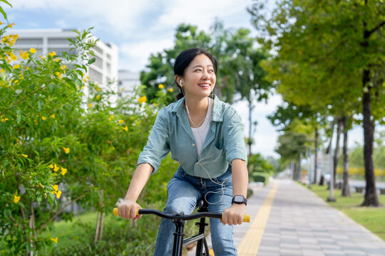 Beautiful Asian woman smiling and listening to her favorite music while cycling on a road in a nature park. Recreational activity. Nature therapy.