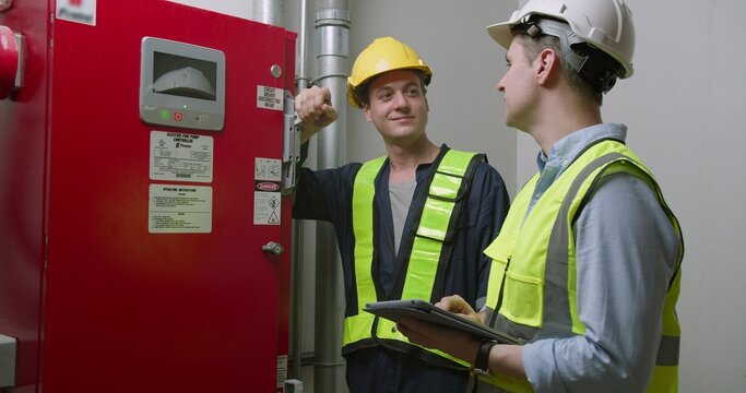 Two engineers collaborate on an industrial safety inspection, using a digital tablet to monitor a fire pump controller system in a facility's mechanical room.