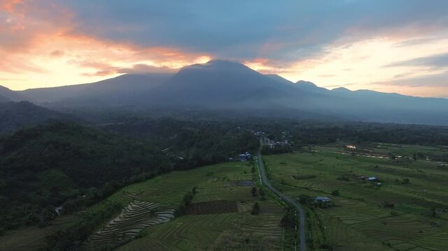 Aerial view of lush rice terraces and a winding road beneath a majestic mountain at sunset in Kemumu, Arga Makmur, Bengkulu, Indonesia.