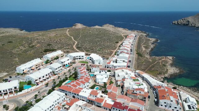 Aerial view of white buildings of Fornells village along the coastline with turquoise waters and rocky cliffs in Fornells, Balearic Islands, Spain.