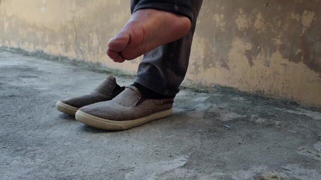 Someone is putting on shoes and socks while leaning against a worn-looking wall. A pair of scuffed brown slip-on shoes sits beside their feet. The concrete floor shows signs of age and wear.