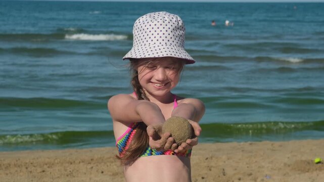Smiling girl in panama hat holding sand ball on beach. Concept of kids summer seaside activities
