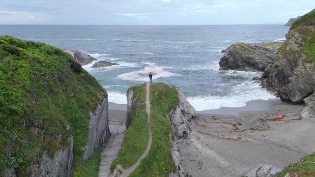 Aerial view of a person standing on a grassy cliff edge overlooking the sandy shore and rocky coastline of Lansallos Beach in Looe, Cornwall, United Kingdom.