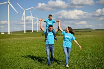 Obraz premium Family walks in green field near wind turbines. Father carries child on shoulders, mother holds hand. Everyone wears blue volunteer shirts. Sunny day, blue sky, white clouds. Happy mood.
