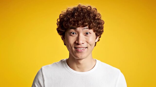 Surprised Young Asian Man with Curly Hair Covering Mouth in Shock on Yellow Background, ideal for reaction content and emotional marketing.