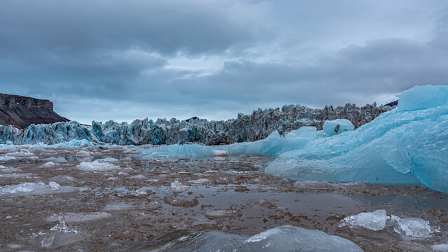eisblaue Eisschollen treiben im schokobraunen Meer vor der Gletscherkante des Kongsbreen in Spitzbergen, dar&uuml;ber bew&ouml;lkter Himmel, Wildnis pur im arktischen Ozean