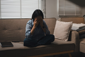 A stressed woman sitting on a couch covering her face with hands in a living room © PRIME STOCK LAB