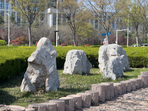 Urban park landscape with decorative rocks, manicured hedges, pebble pathway and spring trees under bright daytime light beside modern buildings creating calm public green space