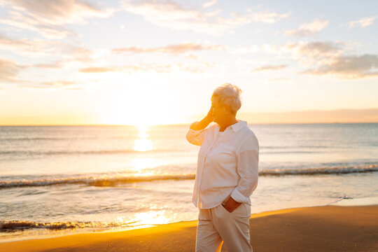 Portrait of gray-haired woman strolling along beach at sunset. Elderly woman enjoys seascape and feels freedom of open air. Concept of happy old age, relaxation, and freedom.