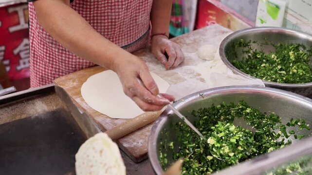 An adult female cook prepares dough with chopped vegetable filling in a kitchen workspace, using a rolling pin.