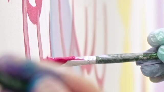 A gloved hand expertly applies bright pink paint to a white canvas. Wavy, flowing lines of fuchsia pigment create an abstract design, adding vibrant color to the evolving artwork in an artist studio.