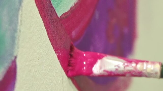A close-up view of a brush gliding across a canvas, applying thick, vibrant magenta paint. The artist creates striking lines, layering the bold color over purple and green elements.