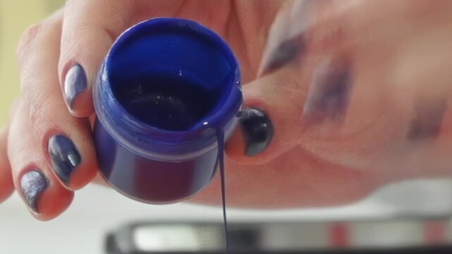 A close-up reveals hands with shimmering nails gently tilting a small blue pot. A mesmerizing stream of dark blue liquid spills out, hinting at artistic endeavors in a bright workspace.