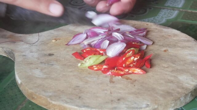 A person's hands are chopping shallots and red chilies on a traditional wooden chopping board.