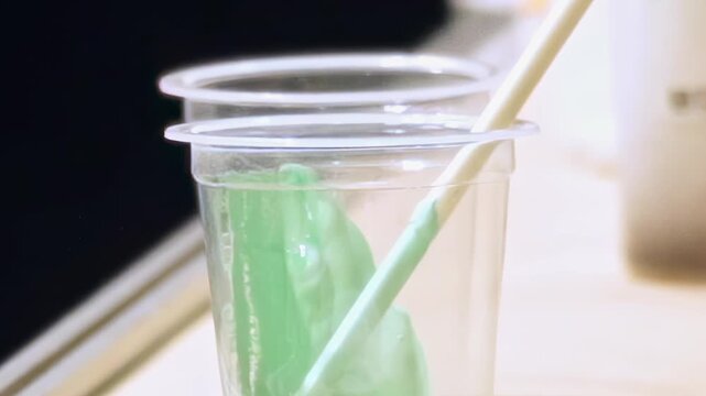 Close-up view reveals clear cup holding brilliant green epoxy resin. Wooden stick gently stirs thick liquid, blending components for creative art piece on bright craft table during day.
